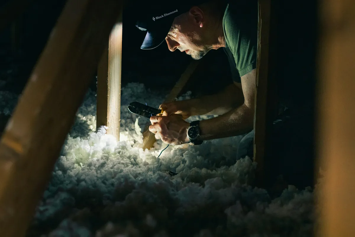 A worker in a dark attic lit by a headlamp, kneeling between exposed roof rafters