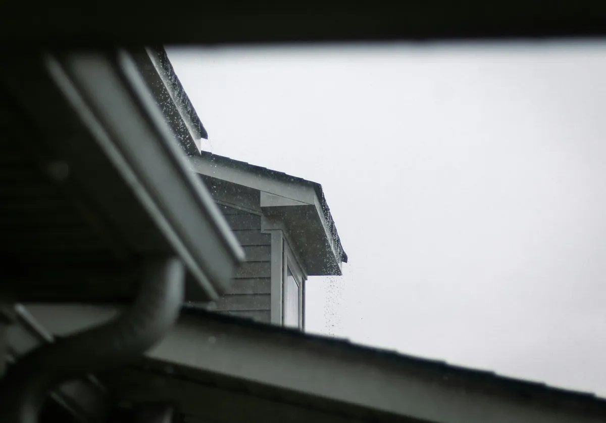 Rain falling past a house gutter and downspout, with a grey sided gable under an overcast sky