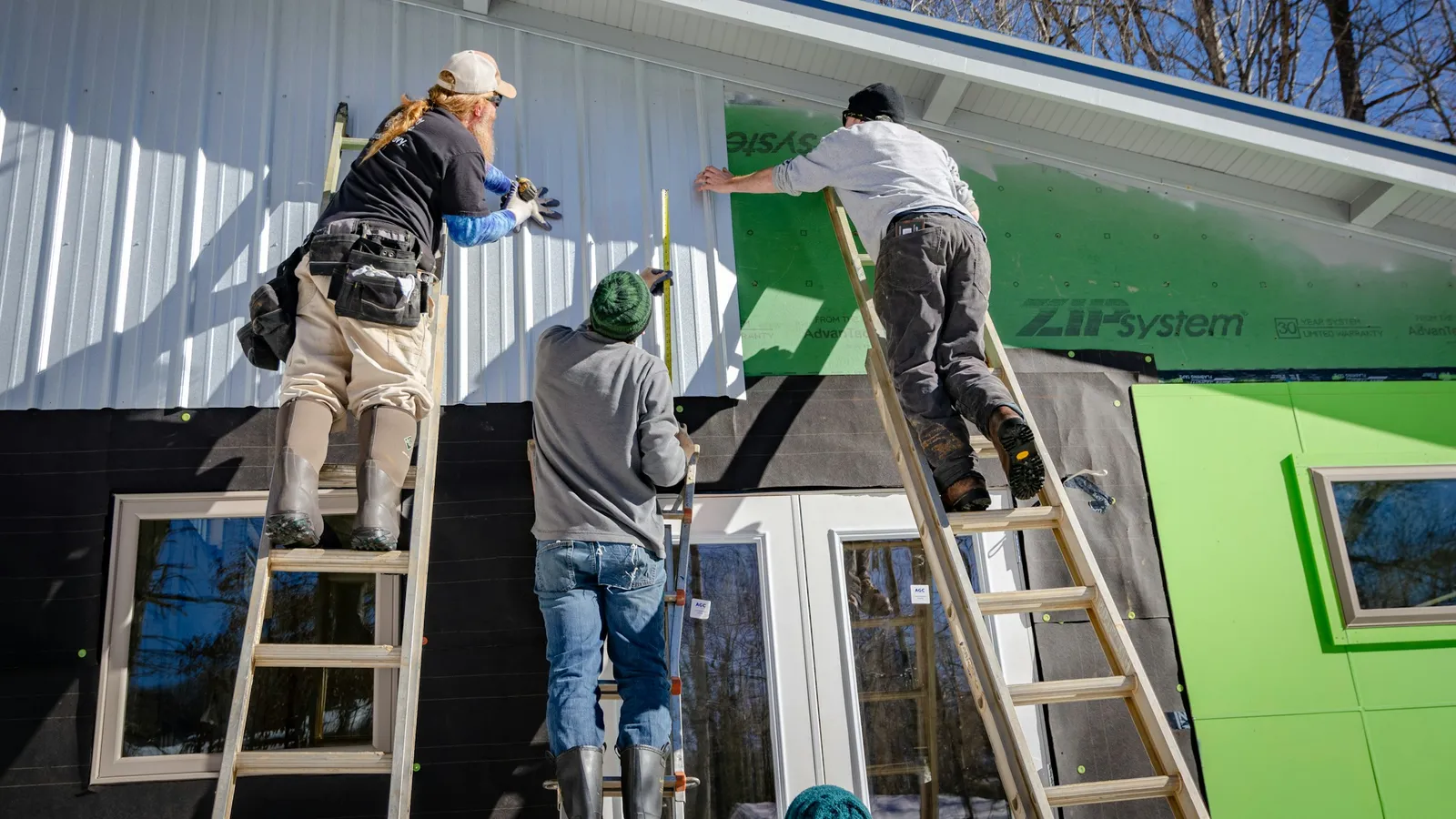 Three workers on ladders fitting metal siding over taped green sheathing panels