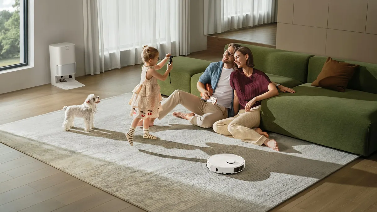 A robot vacuum working a living room rug beside its self-emptying base station while a family sits on the sofa