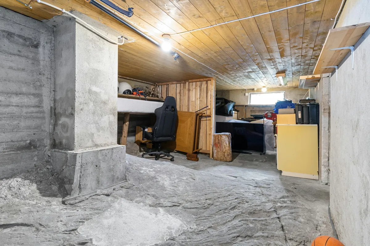 Low unfinished room with bare poured concrete walls, a wooden plank ceiling and stored furniture