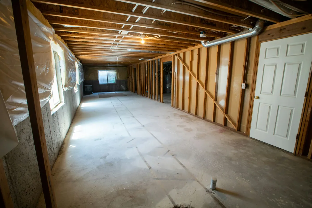 Unfinished basement with a framed stud wall, plastic sheeting on the foundation and a bare concrete floor