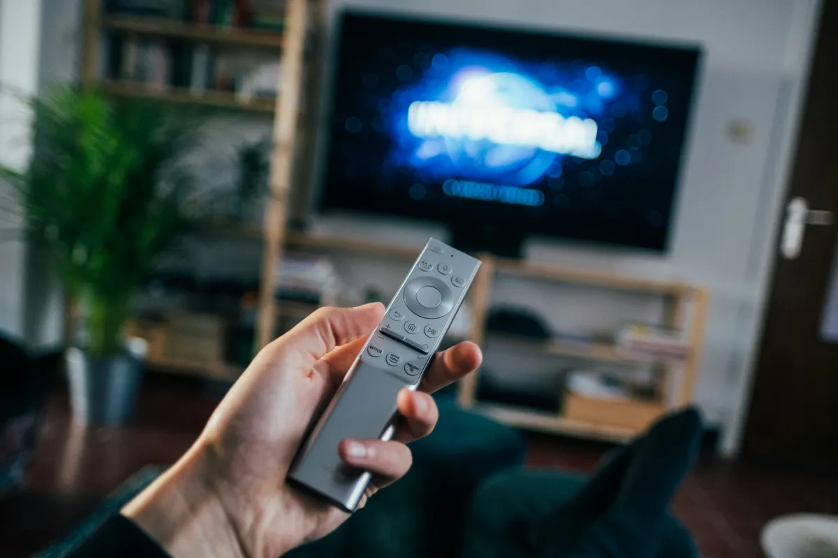 Hand holding a grey remote in front of a television standing on a shelf