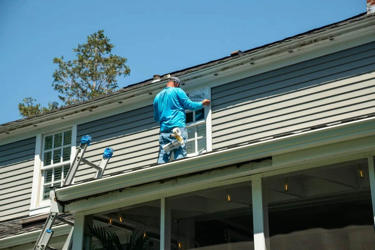 A worker on a ladder pressing tape around an upper-storey window frame on a sided house