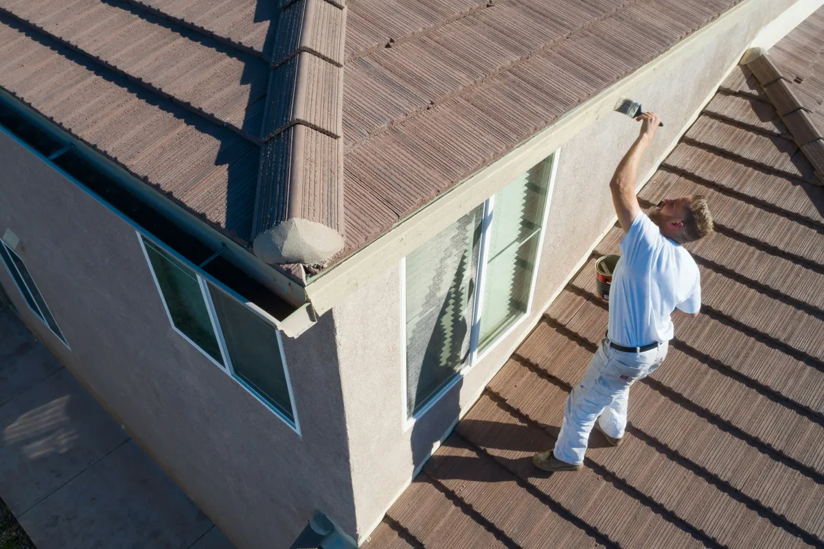 A painter on a roof brushing fresh paint onto a house fascia board