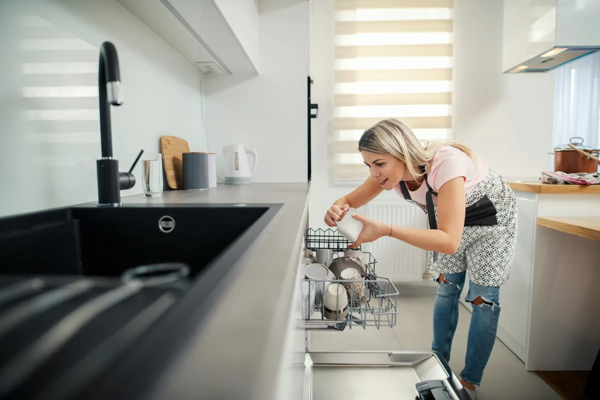A person loading cups into the lower basket of a dishwasher in a bright kitchen